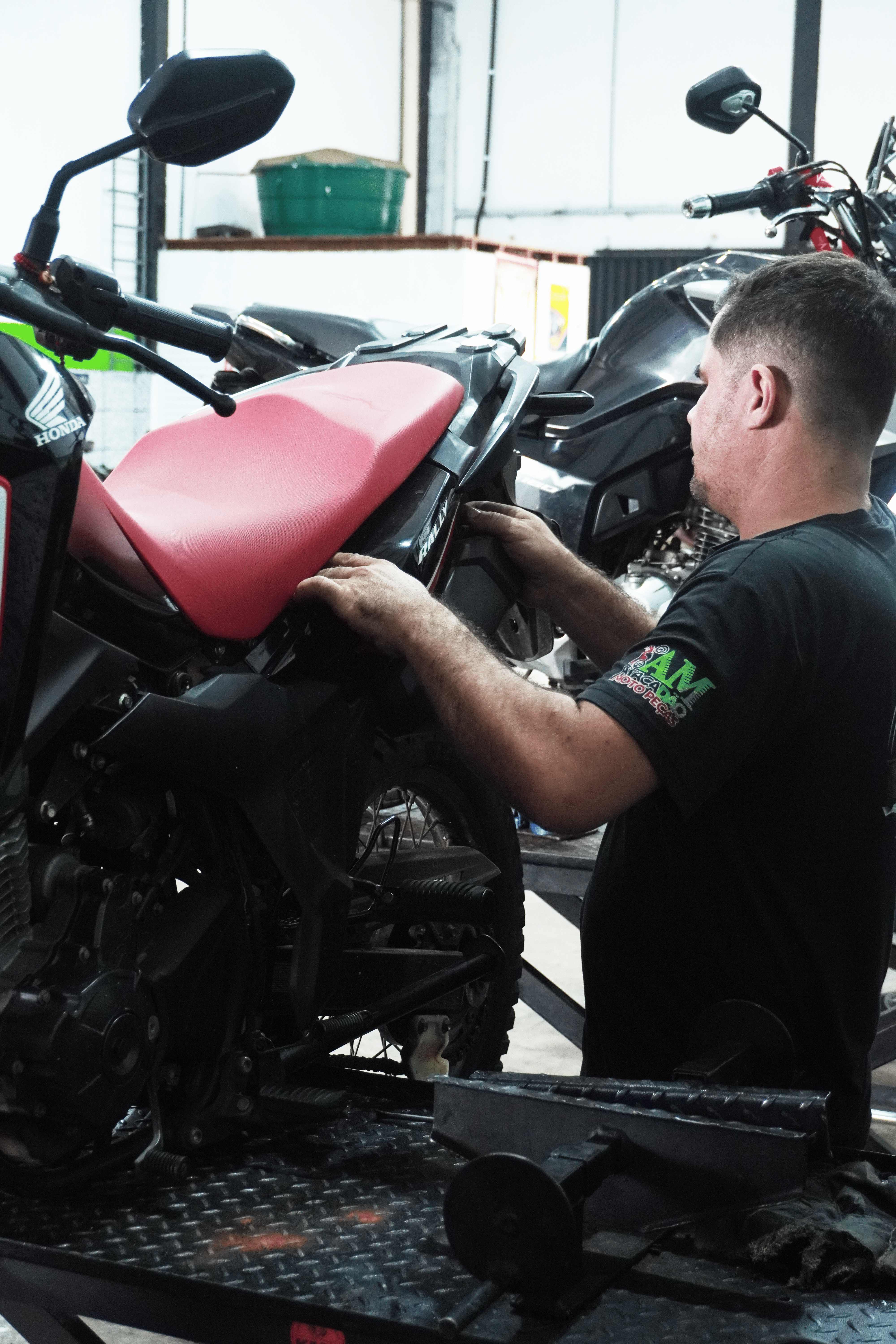 Mecânico ajustando peça de motocicleta com jaqueta vestindo a camisa da Atacadão Moto Peças